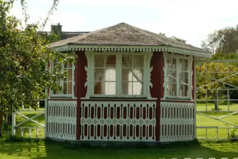 Pavillon de jardin rouge et blanc, orné de dentelle de bois. Une pomme tombe d'un arbre, peut-être pour échapper à la taxe abri de jardin.