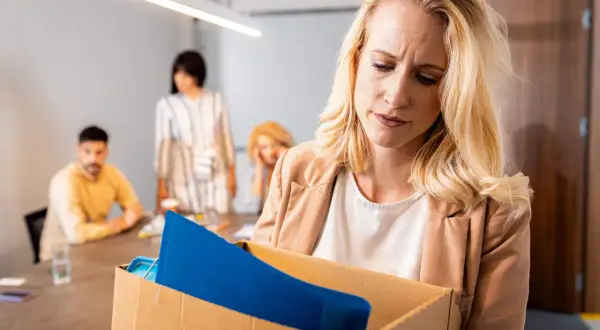 Femme blonde triste portant une boîte de ses affaires, se demandant si l'on peut licencier sans motif. Des collègues flous en arrière-plan.