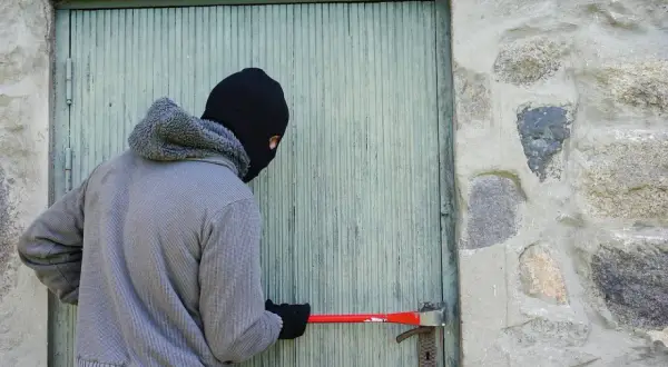 Un individu masqué tente de forcer une porte avec un pied-de-biche, acte relevant du cambriolage et du code pénal.