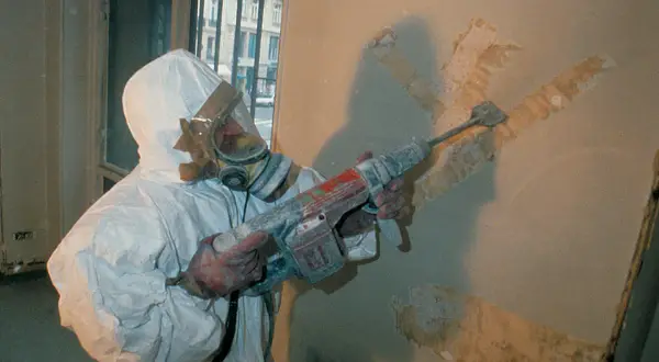 Travailleur en combinaison intégrale et masque à gaz, utilisant un marteau-piqueur pour retirer un revêtement mural. Que faire face à l'amiante au travail.